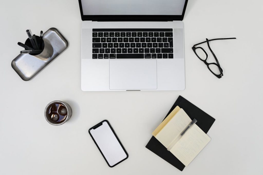 Top view of modern laptop and smartphone with empty screens placed near opened notebook and eyeglasses on light table